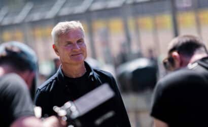 Smiling older man with white hair in a black shirt outdoors, as photographers prepare a shot nearby.