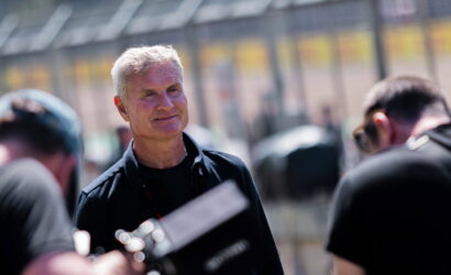 Older man with gray hair smiling, standing in a pit area with blurred figures around him.