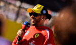 F1 driver in red Ferrari gear wearing a yellow cap and sunglasses, smiling while giving a Sky Sports interview with a microphone labeled Sky Sports in front of him