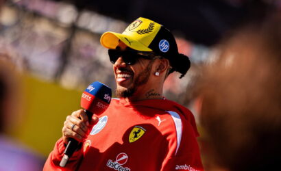 F1 driver in red Ferrari gear wearing a yellow cap and sunglasses, smiling while giving a Sky Sports interview with a microphone labeled Sky Sports in front of him