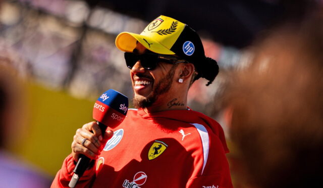 F1 driver in red Ferrari gear wearing a yellow cap and sunglasses, smiling while giving a Sky Sports interview with a microphone labeled Sky Sports in front of him