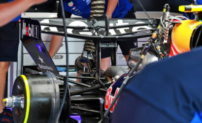 Close-up of a Formula 1 race car in the pit with visible sponsor logos on the rear wing and wheel, mechanics working in the background.