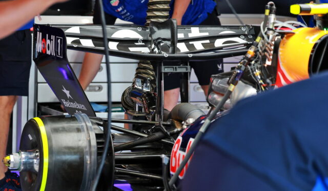 Close-up of a Formula 1 race car in the pit with visible sponsor logos on the rear wing and wheel, mechanics working in the background.