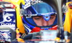 Close-up of a race car driver wearing a blue-visor helmet in the cockpit, sponsor logos visible on gear and carSide tank.