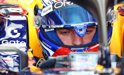 Close-up of a race car driver wearing a blue-visor helmet in the cockpit, sponsor logos visible on gear and carSide tank.