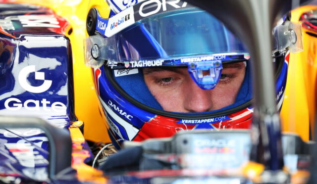 Close-up of a race car driver wearing a blue-visor helmet in the cockpit, sponsor logos visible on gear and carSide tank.
