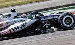 Formula 1 car speeding along a race track with sponsor logos on its white and black livery, driver in helmet visible
