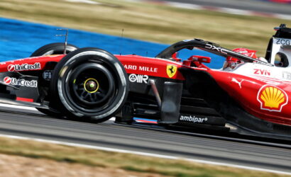 Red Ferrari Formula 1 car on a race track speeding through a corner, with sponsor logos visible on the bodywork, left front wheel in motion.