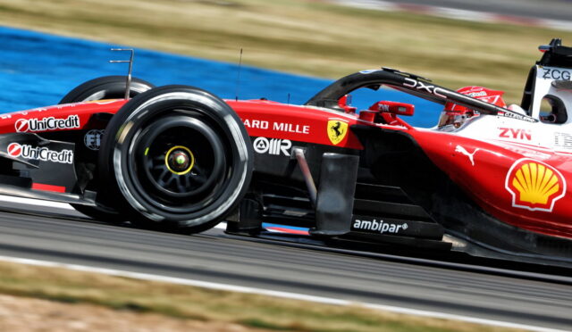 Red Ferrari Formula 1 car on a race track speeding through a corner, with sponsor logos visible on the bodywork, left front wheel in motion.