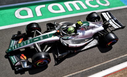 Green‑white Formula 1 car on the track beside an Aramco‑green curb, driver wearing a green helmet, visible sponsor logos on the car.