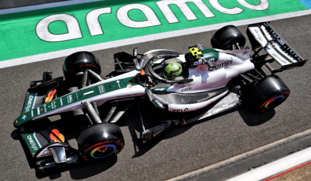 Green‑white Formula 1 car on the track beside an Aramco‑green curb, driver wearing a green helmet, visible sponsor logos on the car.