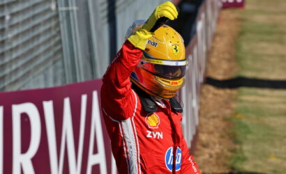 Ferrari race driver in a red suit raises his arm in celebration beside the track, wearing a yellow helmet with sponsor logos.
