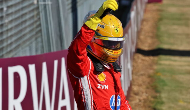 Ferrari race driver in a red suit raises his arm in celebration beside the track, wearing a yellow helmet with sponsor logos.