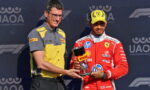 Two men on a podium exchange a trophy; one wears a yellow and gray Pirelli shirt, the other a red racing suit and yellow cap with Ferrari logo.