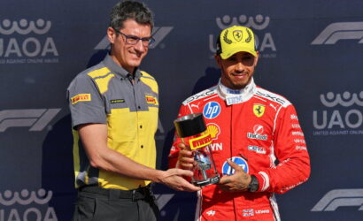 Two men on a podium exchange a trophy; one wears a yellow and gray Pirelli shirt, the other a red racing suit and yellow cap with Ferrari logo.