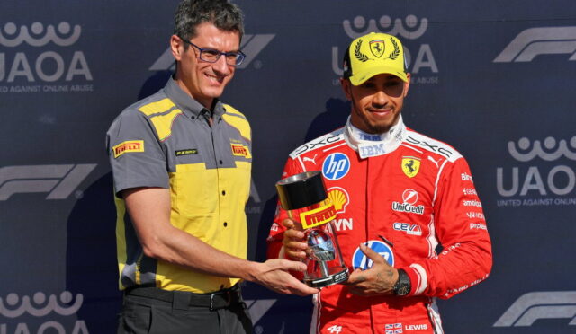 Two men on a podium exchange a trophy; one wears a yellow and gray Pirelli shirt, the other a red racing suit and yellow cap with Ferrari logo.