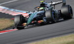 Formula 1 single-seater car on a track taking a tight corner, driver helmet visible in cockpit.