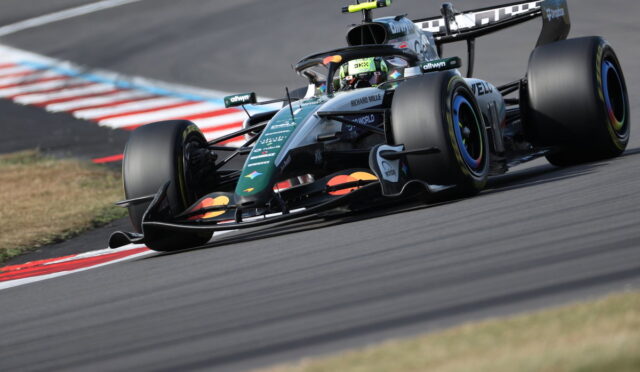 Formula 1 single-seater car on a track taking a tight corner, driver helmet visible in cockpit.