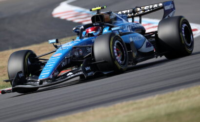 Blue Formula 1 race car taking a tight corner on a race track, with sponsor logos visible along the body and wing.