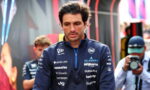 Male race car driver in a navy team jacket with sponsor logos, looking down in a paddock with photographers in the background