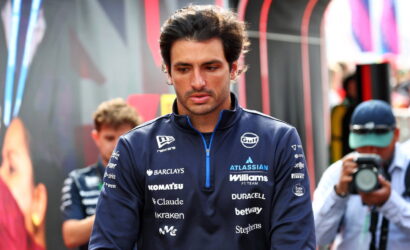 Male race car driver in a navy team jacket with sponsor logos, looking down in a paddock with photographers in the background