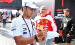 Man in a white cap and sunglasses signs a cap for a fan at a Formula 1 event, with a Ferrari team member in a red jacket behind him in the paddock.