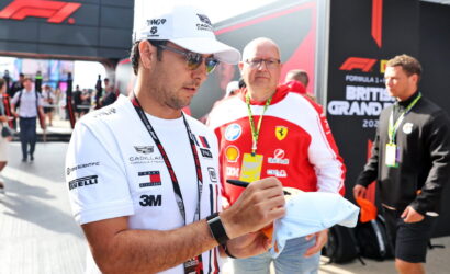 Man in a white cap and sunglasses signs a cap for a fan at a Formula 1 event, with a Ferrari team member in a red jacket behind him in the paddock.