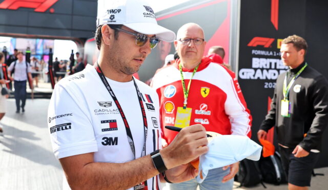 Man in a white cap and sunglasses signs a cap for a fan at a Formula 1 event, with a Ferrari team member in a red jacket behind him in the paddock.
