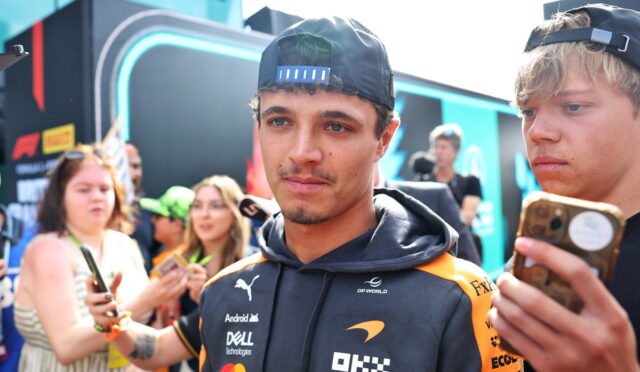 Racing driver in a black and orange DP World jacket, wearing a backward cap, posing while fans take photos nearby.