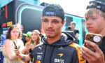 Young man in a branded team jacket with a backward cap, posing among fans at a motorsport event while people take photos nearby