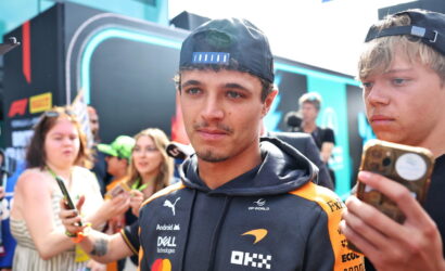 Young man in a branded team jacket with a backward cap, posing among fans at a motorsport event while people take photos nearby