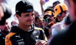 Smiling racer in a black and orange jacket signs autographs as excited fans crowd around him outdoors.