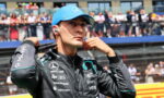 Formula 1 driver in a blue cap and black Petronas racing suit adjusts his earphones before a race, with a cheering crowd behind him.