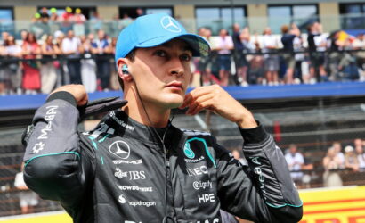 Formula 1 driver in a blue cap and black Petronas racing suit adjusts his earphones before a race, with a cheering crowd behind him.