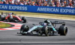 Mercedes Formula One car leads a race on a curved track with red-and-white curbs and a crowded grandstand in the background.