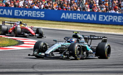 Mercedes Formula One car leads a race on a curved track with red-and-white curbs and a crowded grandstand in the background.