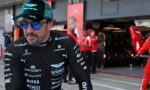 Racing driver in black sponsor-laden suit and green cap walks in a pit lane with red team crew in the background.