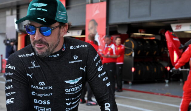 Racing driver in black sponsor-laden suit and green cap walks in a pit lane with red team crew in the background.
