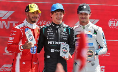 Three race car drivers on a podium holding trophies, wearing racing suits against a red backdrop.