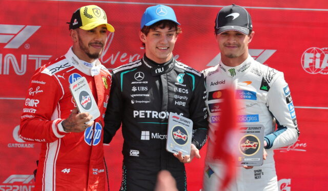 Three race car drivers on a podium holding trophies, wearing racing suits against a red backdrop.