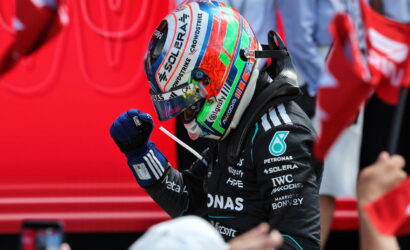 Formula 1 driver in a black Mercedes suit raises a gloved fist, wearing a bright multicolored helmet with sponsor logos; red background.