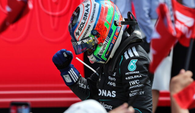 Formula 1 driver in a black Mercedes suit raises a gloved fist, wearing a bright multicolored helmet with sponsor logos; red background.