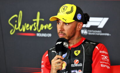 F1 driver in red and black racing suit with a yellow cap, speaking into a microphone during a post-race interview at Silverstone.
