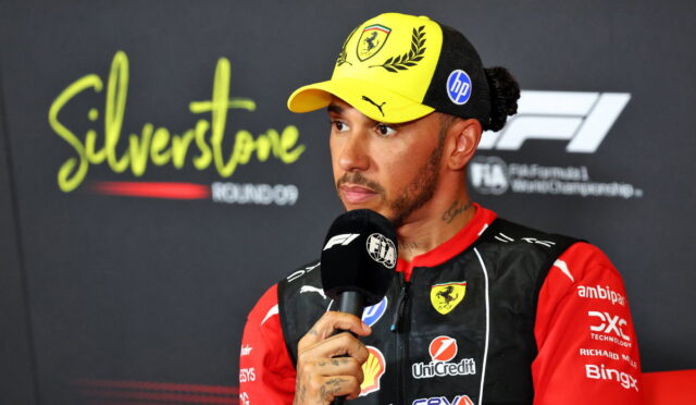 F1 driver in red and black racing suit with a yellow cap, speaking into a microphone during a post-race interview at Silverstone.
