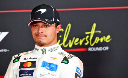 Portrait of a male race car driver in a white sponsor-labeled racing suit and black cap, standing in front of a Silverstone backdrop.