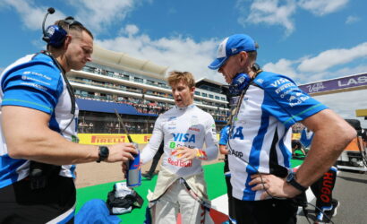 Driver in white racing suit stands between two crew members in blue uniforms at a pit stop, exchanging a Red Bull bottle.