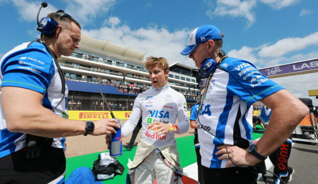 Driver in white racing suit stands between two crew members in blue uniforms at a pit stop, exchanging a Red Bull bottle.