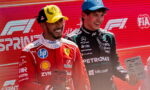 Two Formula 1 drivers pose on a podium, one in red Ferrari gear with a yellow cap, the other in black Mercedes gear with a blue cap, each holding a plaque against a red backdrop with F1 Sprint branding.
