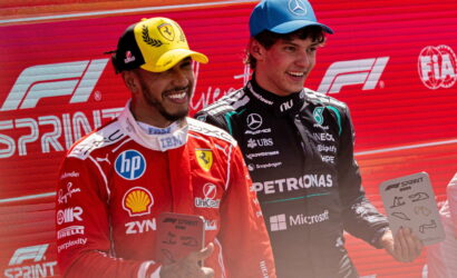 Two Formula 1 drivers pose on a podium, one in red Ferrari gear with a yellow cap, the other in black Mercedes gear with a blue cap, each holding a plaque against a red backdrop with F1 Sprint branding.