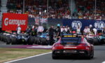Rear view of a red safety car labeled 'CrowdStrike' on a race track, with race cars and a crowded grandstand in the background